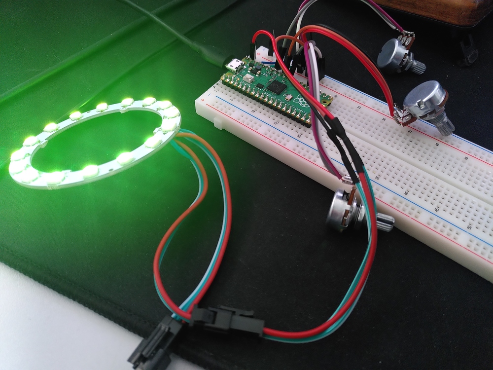 A picture of the prototyped electronics on a breadboard. A Raspberry Pi Pico microcontroller is connected to 3 potentiometers and a 16 LED WS2812 LED ring PCB. The LEDs glow in a bright green.