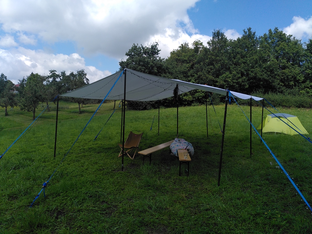 A large white tarp standing on green grass. 2 benches and a chair are underneath the tarp.